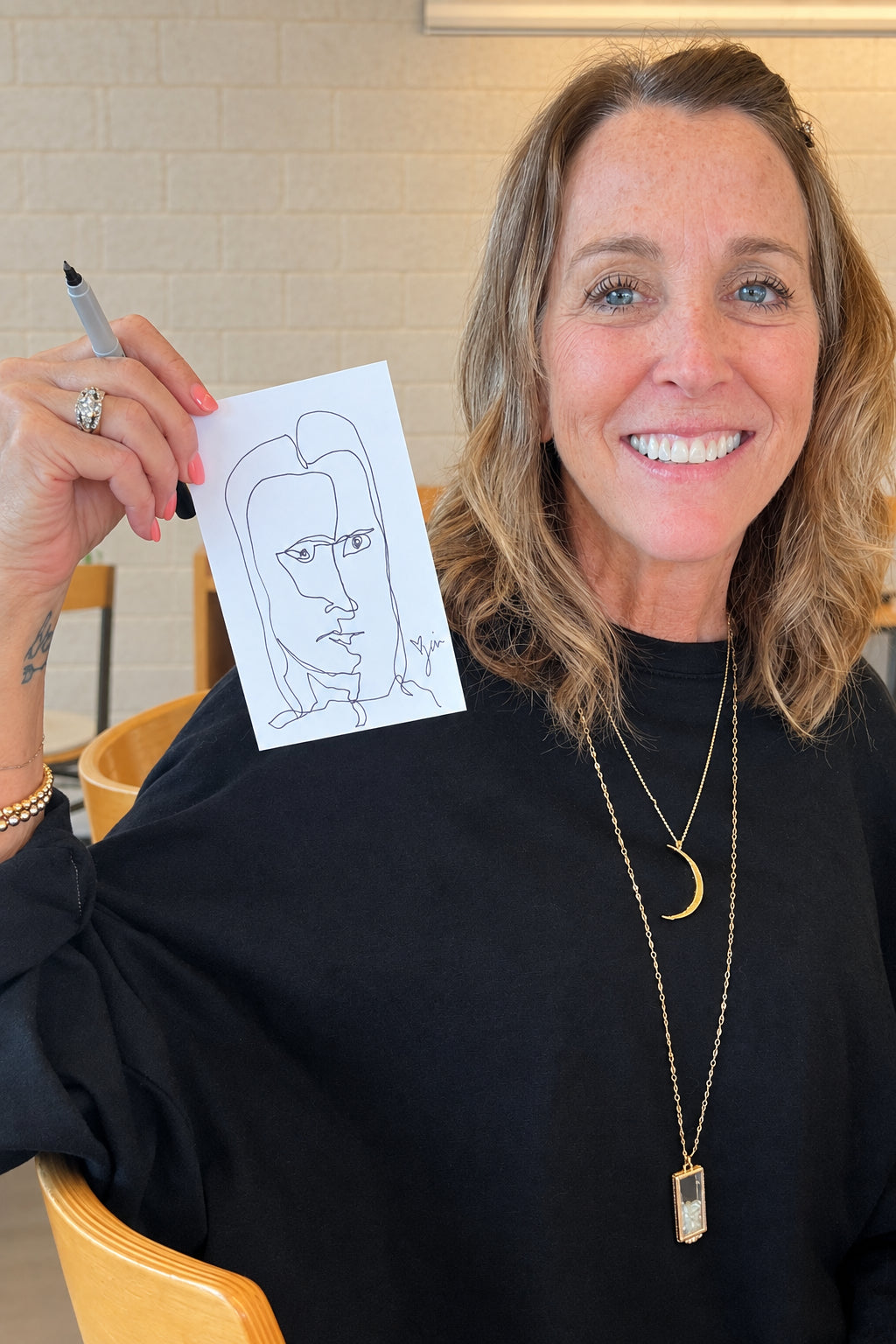 A smiling woman with blonde hair holds up a small white sketch card with a simple line drawing of a face. She is wearing a black top and layered gold necklaces in a bright indoor setting with a brick wall and wooden chairs visible in the background.