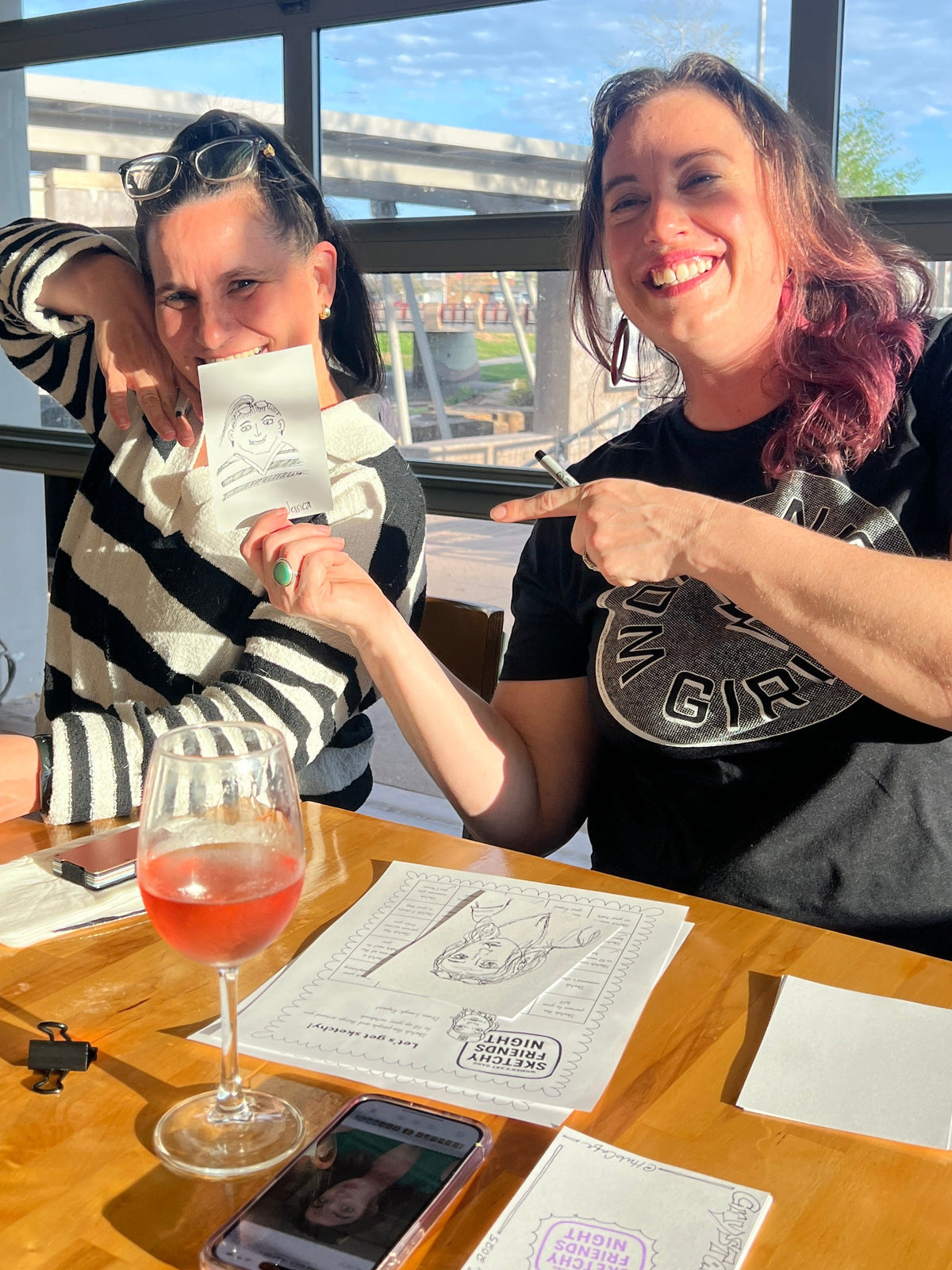 Two women sitting at a wooden table in a restaurant or cafe, laughing and pointing at a printed ticket or receipt. A wine glass, phone, and papers are on the table, with large windows behind them showing an outdoor scene.