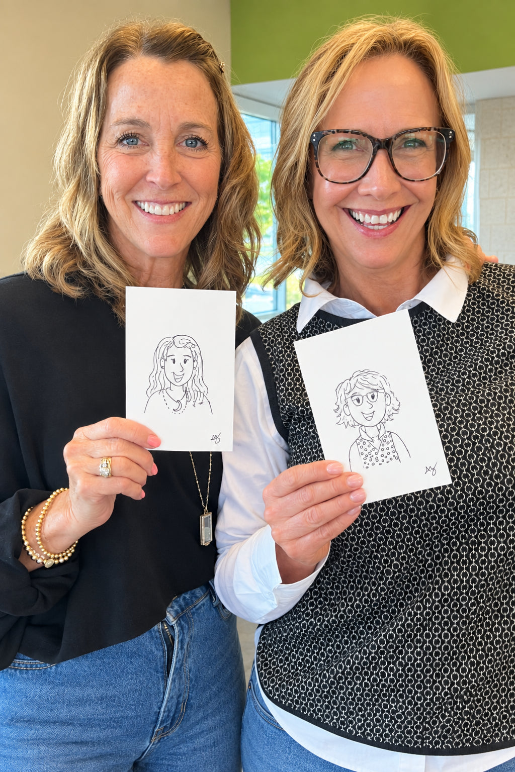 Two smiling women standing indoors and holding up small white sketch cards with simple line drawings. The woman on the left wears a black long-sleeve top and blue jeans, and the woman on the right wears glasses, a white collared shirt, a black-and-white patterned vest, and blue jeans.