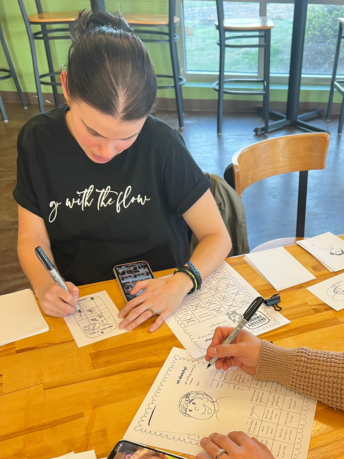 Two people seated at a wooden table in a cafe or studio space, writing on small printed cards or papers with pens. One person wears a black graphic T-shirt, and the other is in a beige knit sweater.