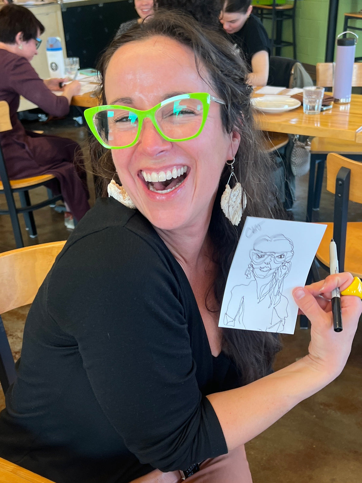 A woman with long dark hair, bright green glasses, and a black top smiles while holding up a small sketch she drew with a black marker. She is seated at a wooden table in a cafe or studio space with other people and tables visible in the background.