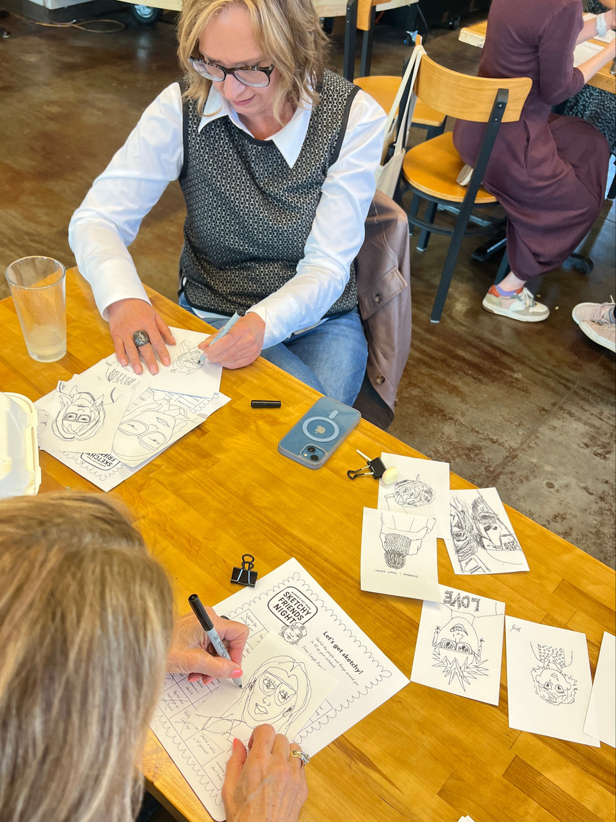 Two women seated at a wooden table in a cafe or studio setting, drawing and writing on printed sheets with pens. Papers, a glass, binder clips, and a phone are scattered on the table.