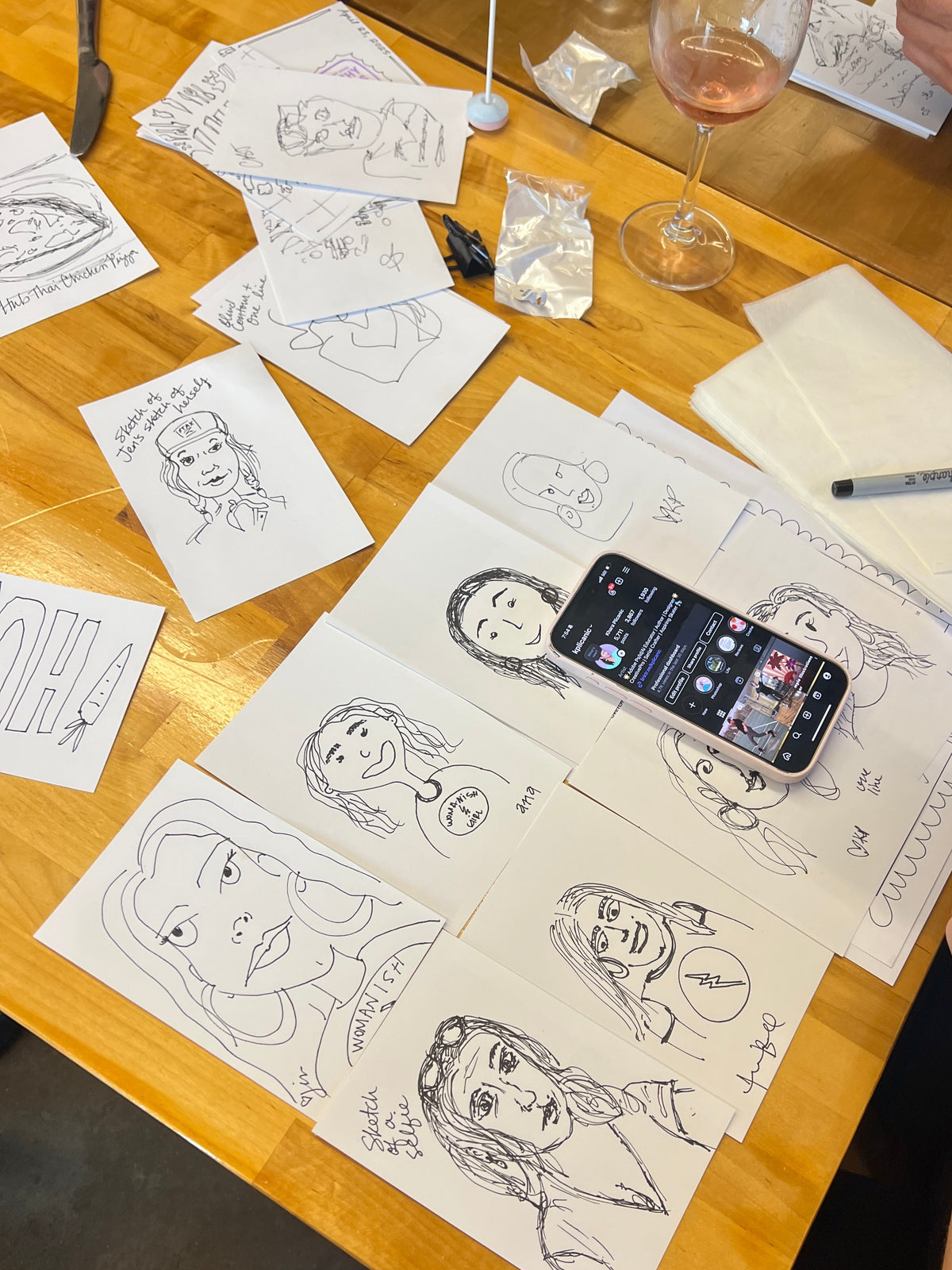 A wooden table covered with hand-drawn black ink caricature portraits on white paper, with a smartphone lying on top and a person's hand holding another phone in the foreground. Papers, a pen, a glass of pink drink, and part of another person are also visible in a casual indoor setting.